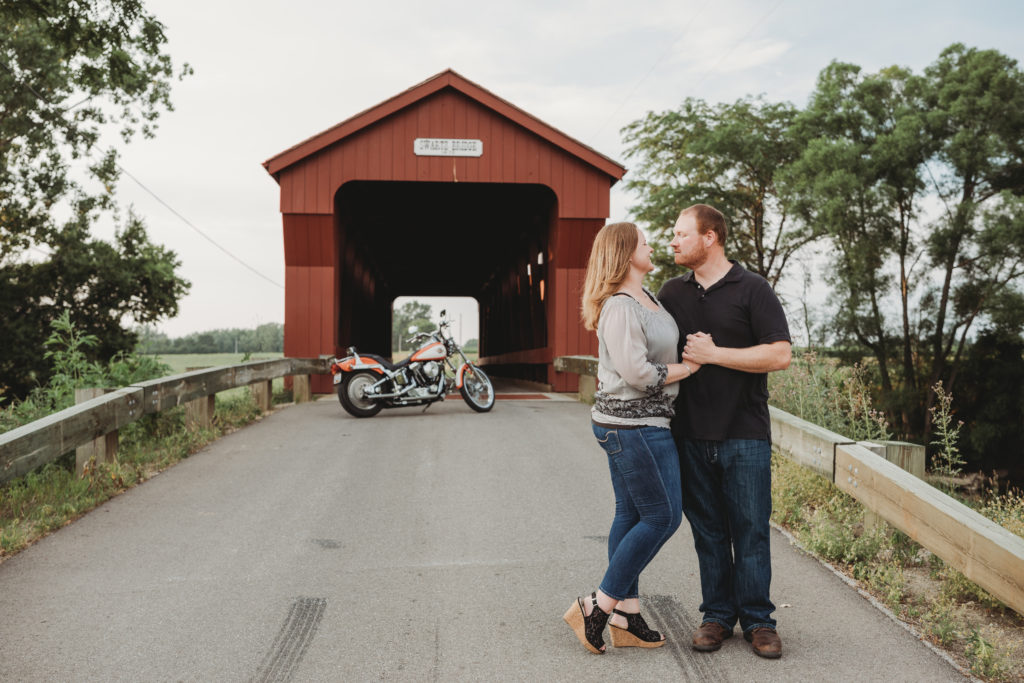 Couples stands together on road in front of the Swartz Covered Bridge. Their motorcycle is parked behind them, by the bridge.