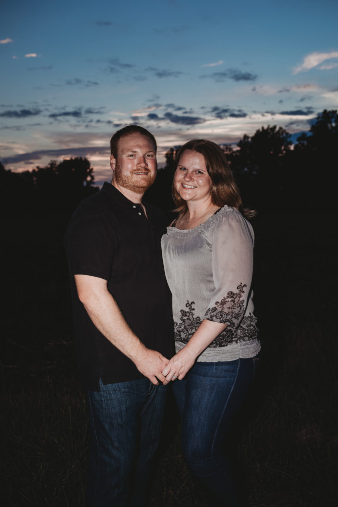 Brandon and Rachel are standing, holding hands, smiling at the camera, with the sunset behind them.