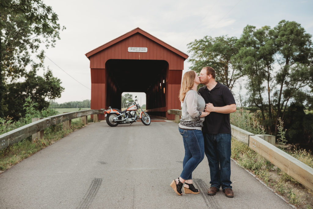 Rachel and Brandon stand together, kissing with his motorcycle parked in the background, close to the entrance of the Swartz Covered Bridge.
