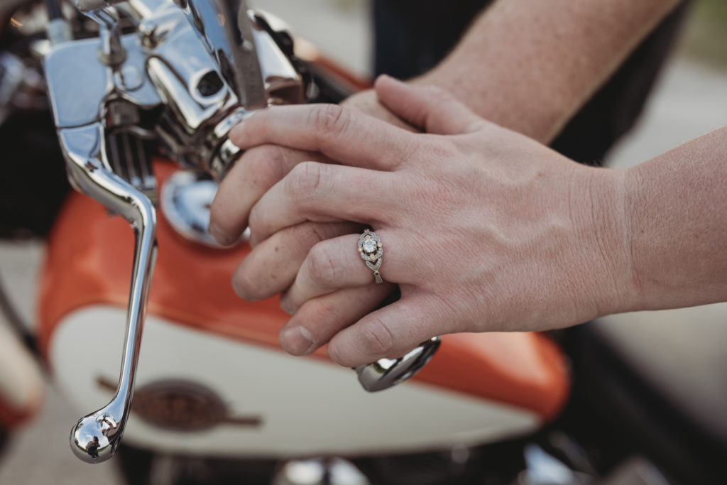 Focus is on Rachel's engagement ring with both their hands on the handle of the motorcycle and a blurred view of the rest of the motorcycle is beyond their hands.