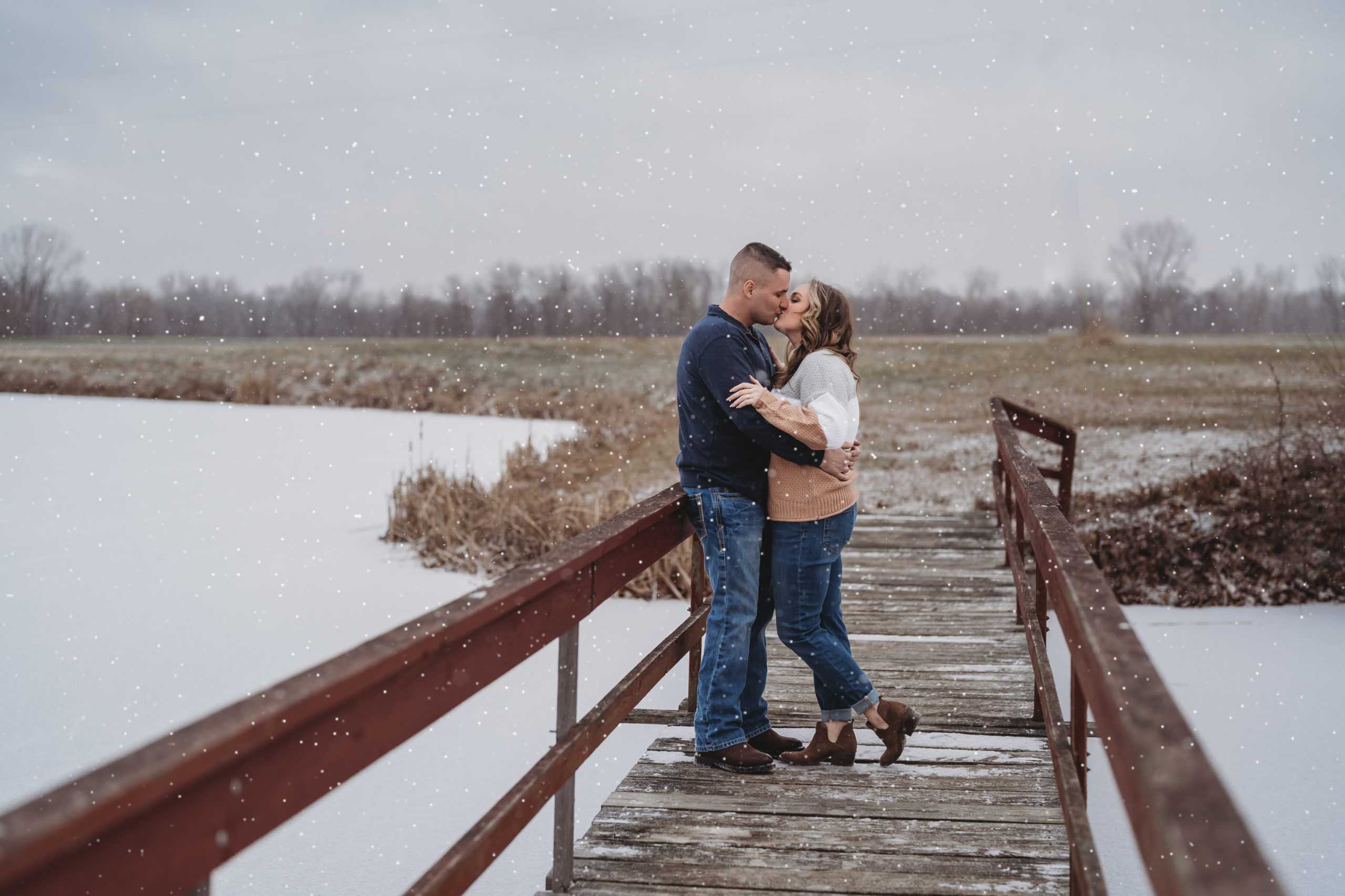 Newly engagement couple embraces one another on bridge over a snow covered pond